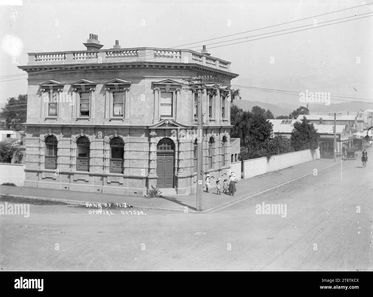 Bank of New Zealand, Opotiki, Blick auf die Bank of New Zealand an der Ecke Church Street und Elliot Street. Eine Gruppe von Frauen und Kindern steht vor der Bank. Auf der rechten Seite wird ein Pferd die Straße hinaufgeritten. Anfang der 1900er Jahre Stockfoto