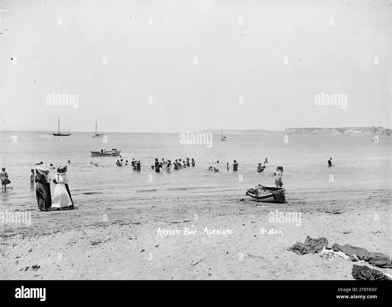 Badegäste in Arkles Bay, Auckland, Neuseeland, Blick auf Erwachsene und Kinder, die im Wasser in der Nähe des Ufers in Arkles Bay spielen. Zwei Frauen stehen am Wasser und beobachten die Badenden. Entworfene Kleidung kann man im unmittelbaren Vordergrund und im Schlauchboot am Wasserrand sehen. Die Küste der East Coast Buys und die Insel Rangitoto sind in der Ferne zu sehen. Aufgenommen in den frühen 1900er Jahren Stockfoto