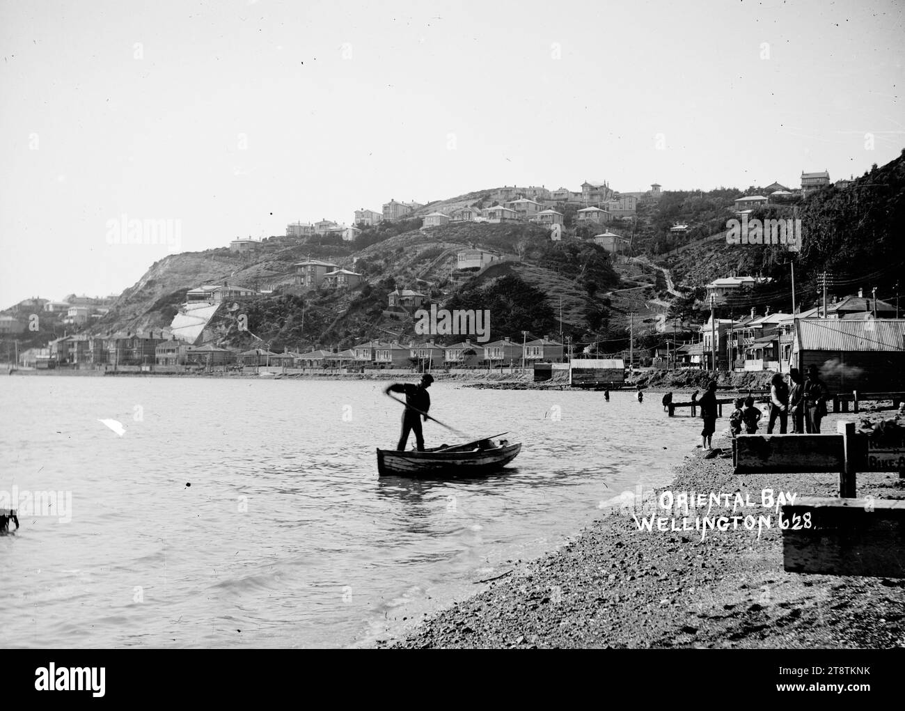 Oriental Bay, Wellington, Neuseeland, Blick auf die Oriental Bay vom Strand mit Blick nach Osten über die Bucht zu den Häusern auf dem Hügel mit Blick auf die Bucht. Es gibt mehrere Bootshäuser am Ufer und Häuser der Oriental Parade. Im Vordergrund steht ein Mann in einem Ruderboot in der Nähe des Ufers. Männer und Jungen sind am Strand versammelt und beobachten ihn. Anfang der 1900er Jahre Stockfoto