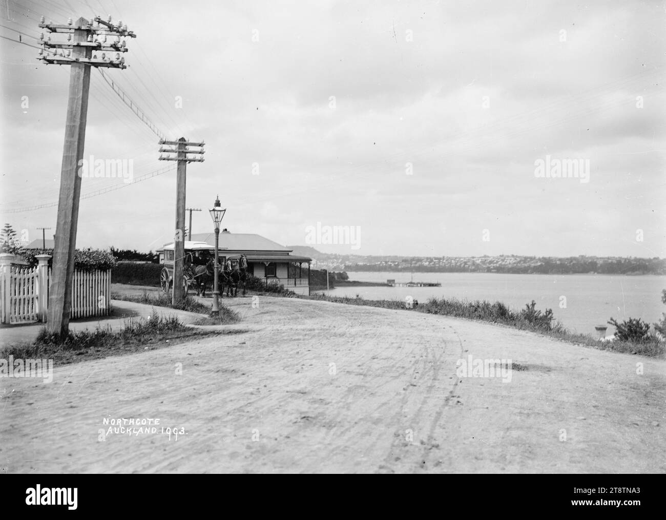 Blick auf die Straße in Northcote, Auckland, Neuseeland, Blick nach Süden in Richtung Northcote Wharf mit Auckland, Neuseeland in der Ferne. Auf der linken Seite befindet sich ein Haus mit Blick auf Little Shoal Bay und den Waitemata Harbour. Auf der linken Seite sind auch ein weißer Pfostenzaun, Stromleitungen und eine Gaslampe zu sehen. Ein Pferdebus befindet sich auf der unbefestigten Straße vor dem Haus. Aufgenommen um 1910 von Auckland, Neuseeland Stockfoto