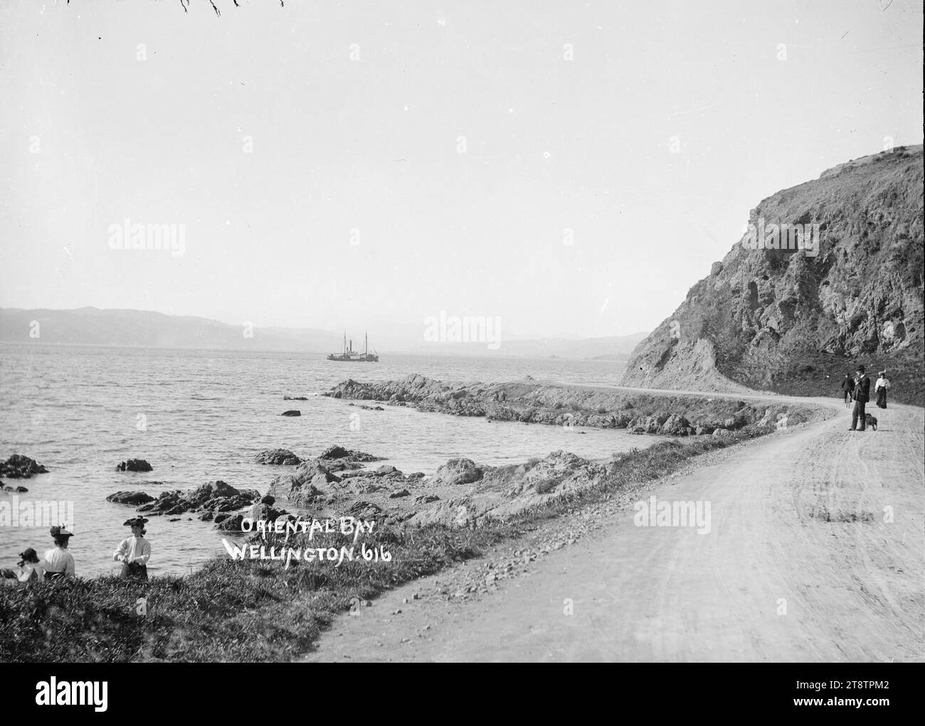 Oriental Bay, Wellington, Neuseeland, Blick auf Lambton Harbour von der Oriental Parade aus in Richtung Nordosten in Richtung Petone mit Somes Island in der Ferne. Ein Fischerboot ? Geht auf den Weg ins Meer. Zwei Frauen und ein Junge stehen auf den Felsen am Ufer im Vordergrund links. Mehrere Leute sind auf der Straße, einer fährt mit dem Fahrrad. Foto zwischen 1910 und 1930 Stockfoto