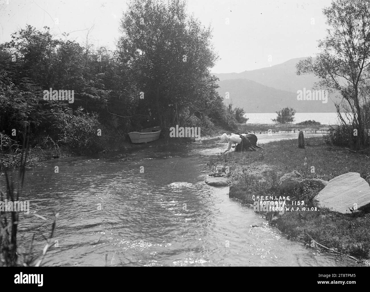 Frauen am Rotokakahi-See, Blick auf zwei Frauen an einem Fluss, der in den Rotokakahi-See mündet. Die Frauen knieten am Wasserrand nieder und sahen etwas im Wasser an. Auf der gegenüberliegenden Seite des Baches, zwischen Bäumen am Wasserrand, befindet sich ein Schlauchboot. Der See ist in der Ferne zu sehen. Foto aufgenommen im Januar 1908 Stockfoto