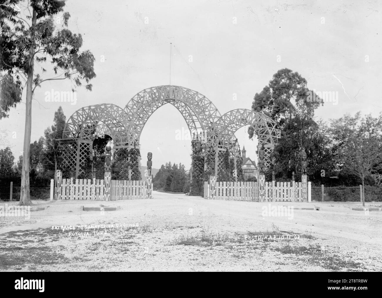 Princes Gate am Eingang zum Gelände des Government Sanatorium and Baths in Rotorua, Blick auf den dreifachen Torbogen und Fahnenmast (bekannt als Princes Gate) am Eingang, der von der Hinemaru Street zum Queens Drive und zu den Gärten des Government Sanatorium and Baths führt. Auf beiden Seiten des Eingangs befindet sich ein hölzerner Zaun aus verzierten Palmen mit geschnitzten Maori-Figuren auf den Zaunpfosten. Das Badehaus Tudor Towers (erbaut 1906-1907) ist in der Entfernung hinter dem Torbogen auf der rechten Seite zu sehen., 10. November 1908 Stockfoto