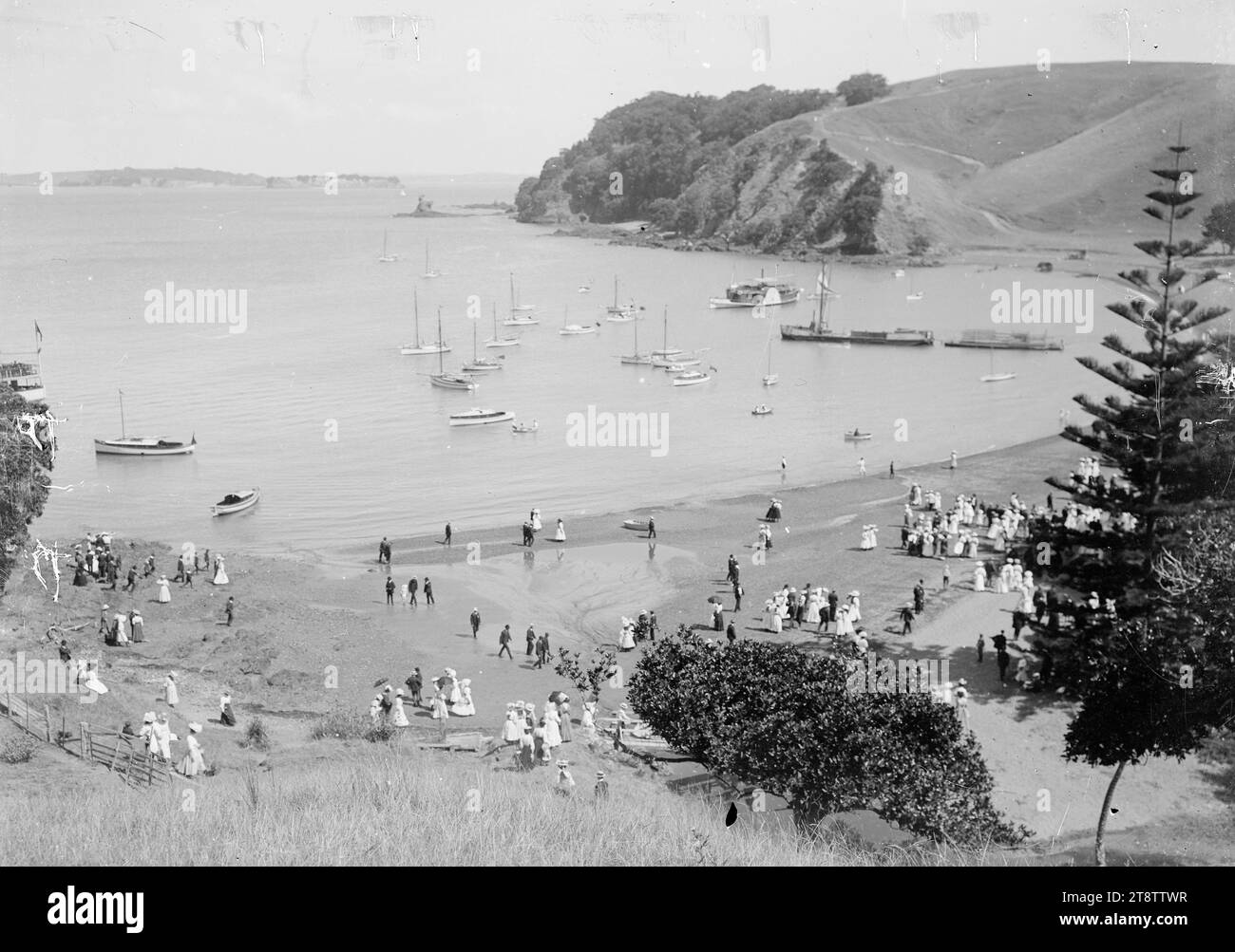 Home Bay, Motutapu Island, Foto um 1910 von William Price. Allgemeiner Blick auf Home Bay, Motutapu Island, von einem Hügel am nördlichen Ende des Strandes aus, der nach Süden in Richtung Motuihe Island in der Ferne blickt. Es gibt viele Boote in der Bucht, einschließlich einer Fähre, und Menschenmassen am Strand Stockfoto