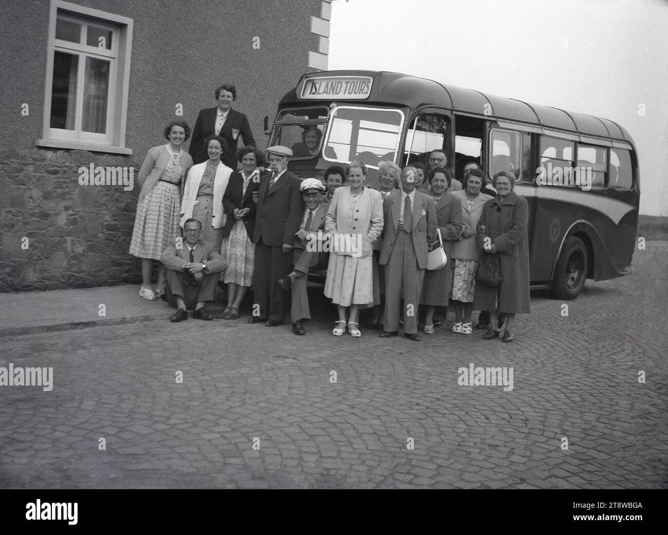 1950er Jahre, historisch, eine Gruppe von Erwachsenen auf einem Ausflug auf Guernsey, Kanalinseln mit ihrem Bus Transport, einem Albion Victor, betrieben von Guernsey Motors. In den formellen Kleidern der Epoche, Männer in Anzügen und Krawatten, Damen in langen Kleidern, versammeln sie sich um den Busfahrer mit seinem Hut, um ein Foto zu machen. Guernsey ist die zweitgrößte der Inseln im englischen Kanal nahe der französischen Küste. Stockfoto