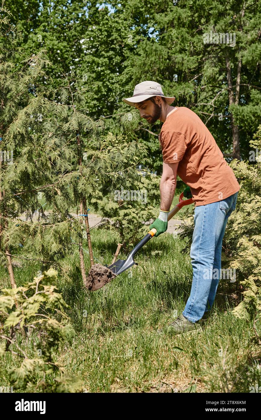 Fröhlicher Gärtner mit Bart und Sonnenhut und Graben mit Schaufel in der Nähe von Pflanzen und Gewächshaus Stockfoto