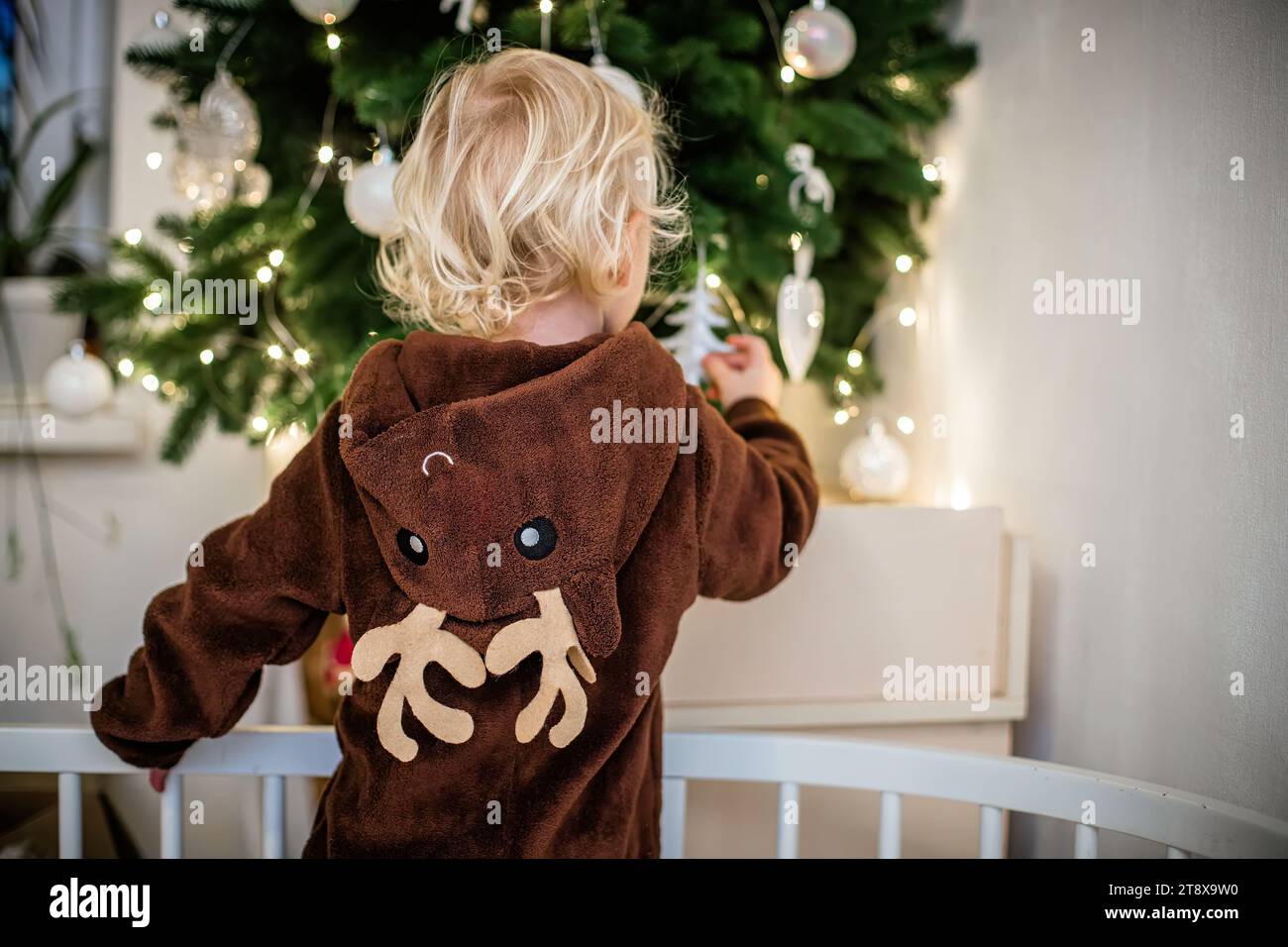 Kleines Mädchen im Rentierkostüm, das den Weihnachtsbaum schmückt, während es in der Krippe steht Stockfoto