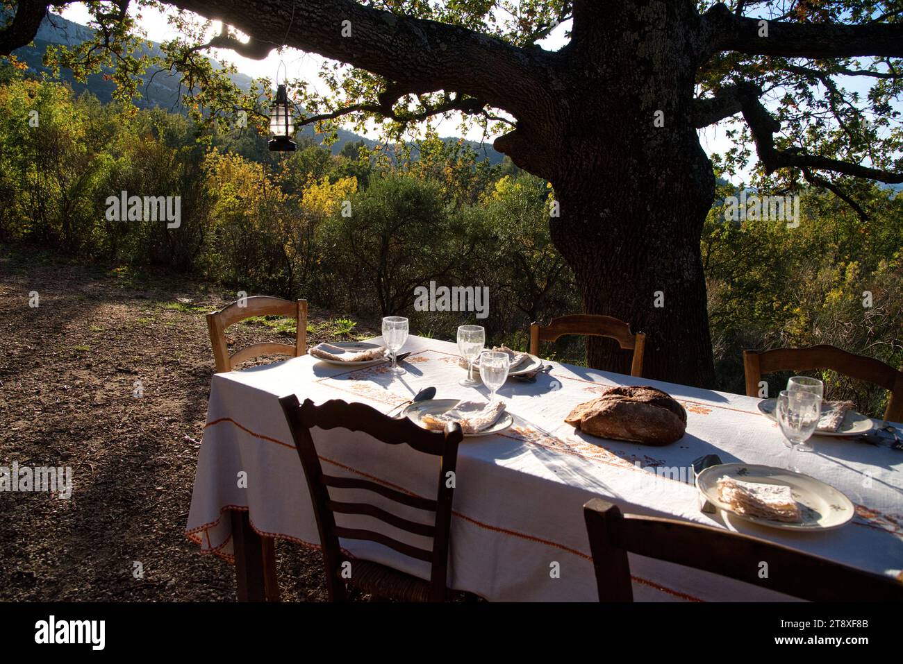 Une table à Manger dressée, avec assiettes, couverts verres et miche de Pain, sur une Terrasse à l'heure du souper, avec en arrière Plan les collines Stockfoto