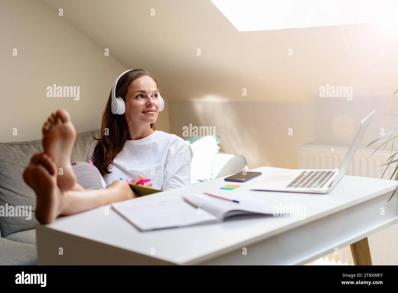 Der Lebensstil des Schülers. Eine junge Frau in weißen Kopfhörern sitzt mit den Füßen auf dem Tisch. Stockfoto