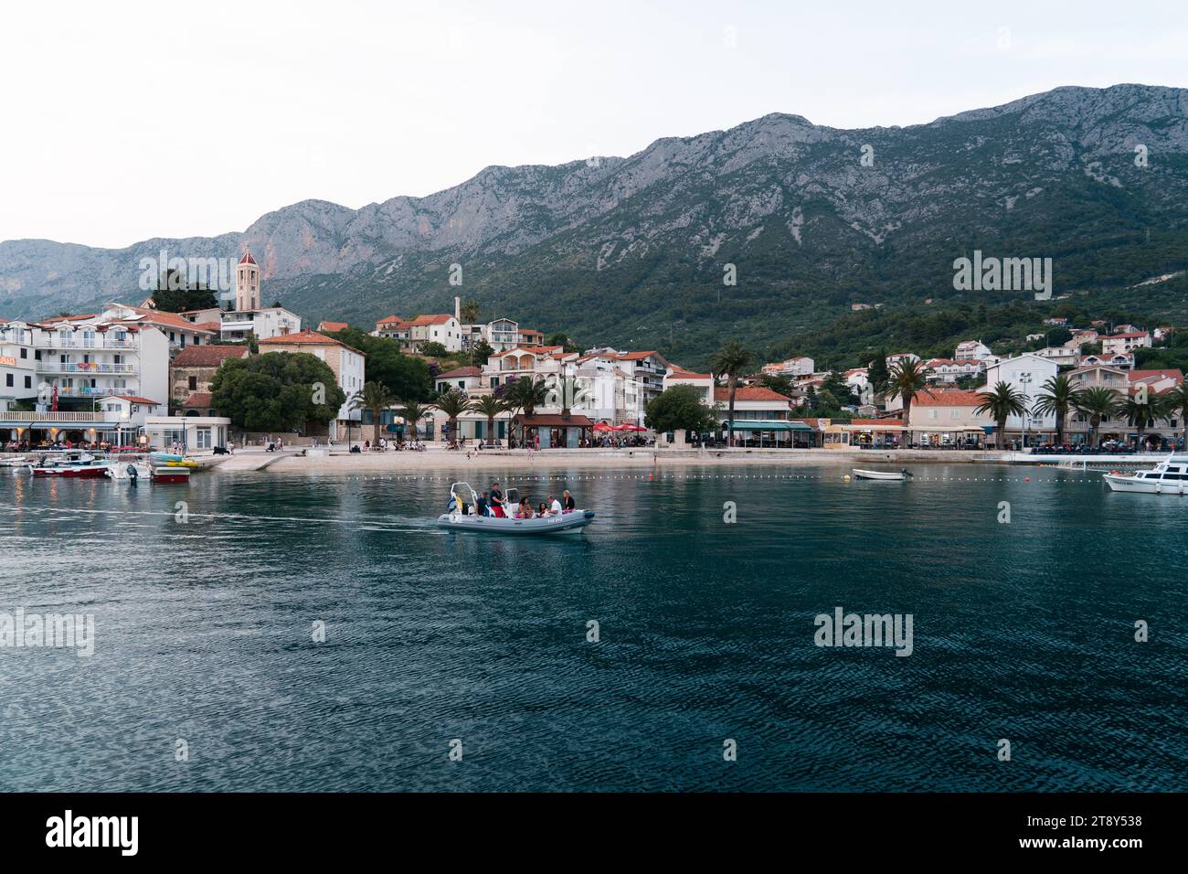 Gradac, Kroatien. Eine Küstenstadt mit einer bergigen Kulisse. Im Vordergrund befindet sich ein kleines Boot mit Menschen auf ruhigem Wasser in der Nähe der Küste. Stockfoto