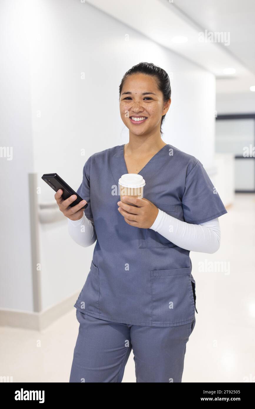 Porträt einer glücklichen birassischen Ärztin mit Smartphone, die im Krankenhaus Kaffee zum Mitnehmen hält Stockfoto