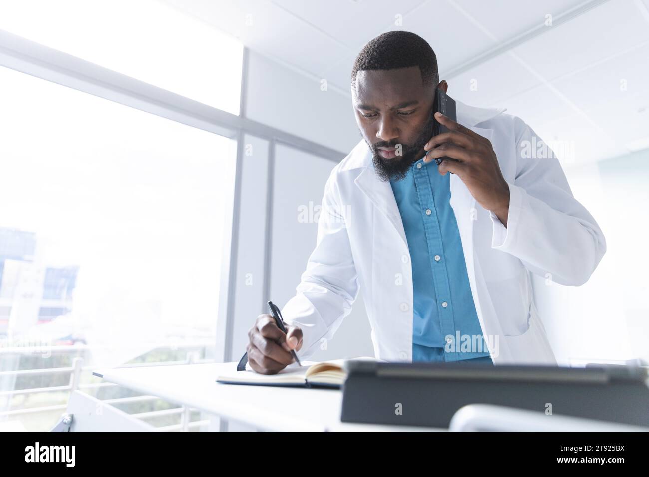 afroamerikanischer männlicher Arzt, der auf dem Smartphone spricht und Notizen im Krankenhauszimmer macht, Kopierraum Stockfoto