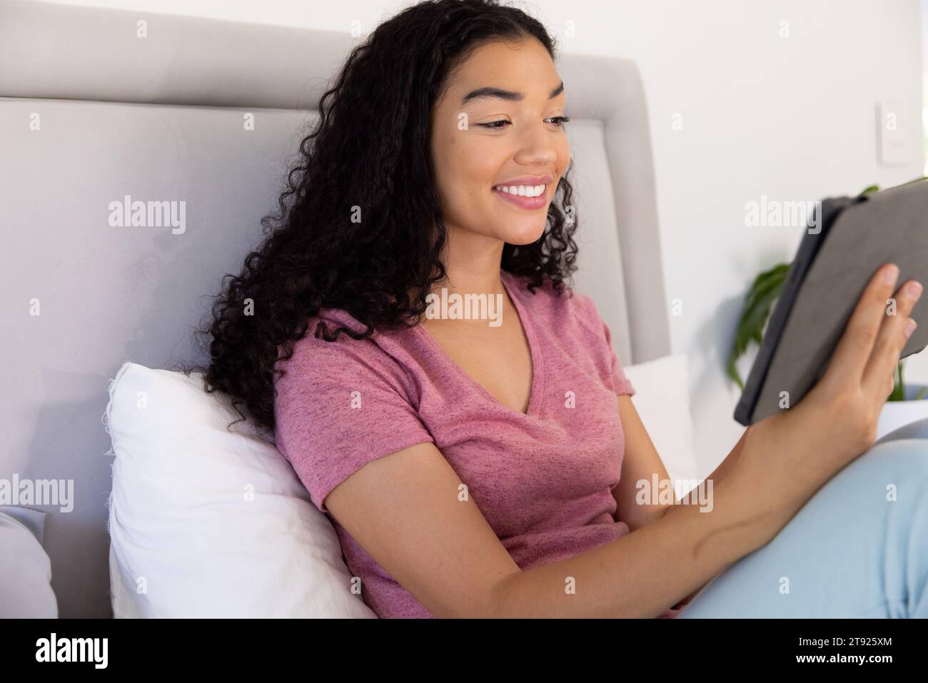 Glückliche birassische Frau, die ein Tablet benutzt, das im sonnigen Haus auf dem Bett sitzt Stockfoto