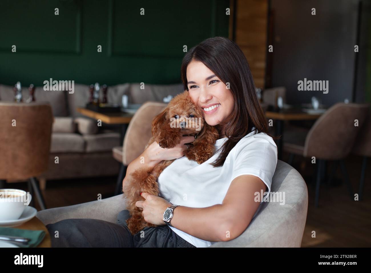 Porträt einer lächelnden Frau, die einen Hund umarmt, während sie am Tisch im Restaurant oder Café sitzt. Beziehungen zwischen Menschen und Haustieren, Liebe und Zuneigung. Stockfoto