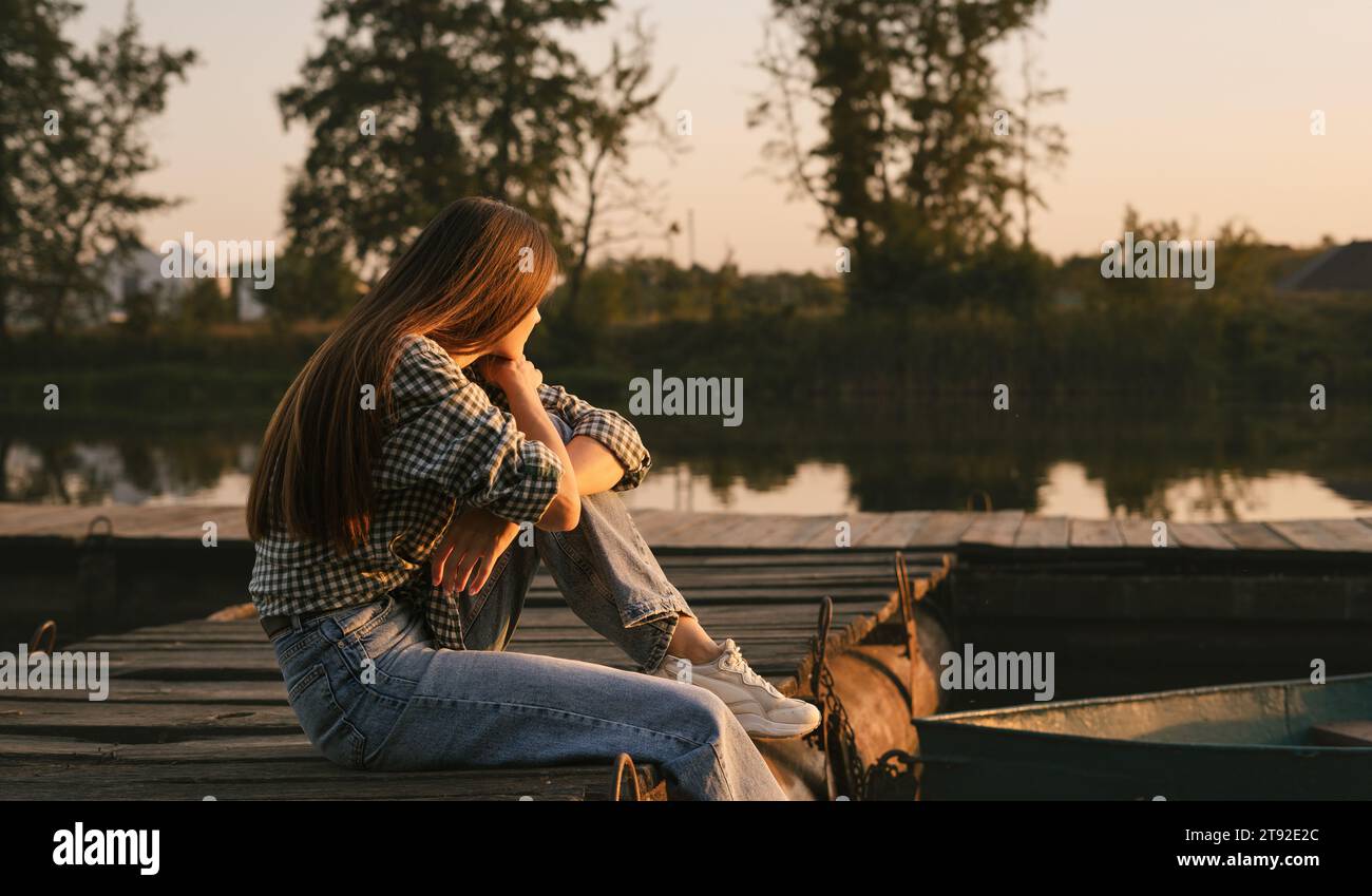 Ein Mädchen mit langen Haaren, das auf dem hölzernen Flussdock sitzt. Es ist Sommer Sonnenuntergang auf dem Fluss. Gutes und warmes Wetter. Stockfoto