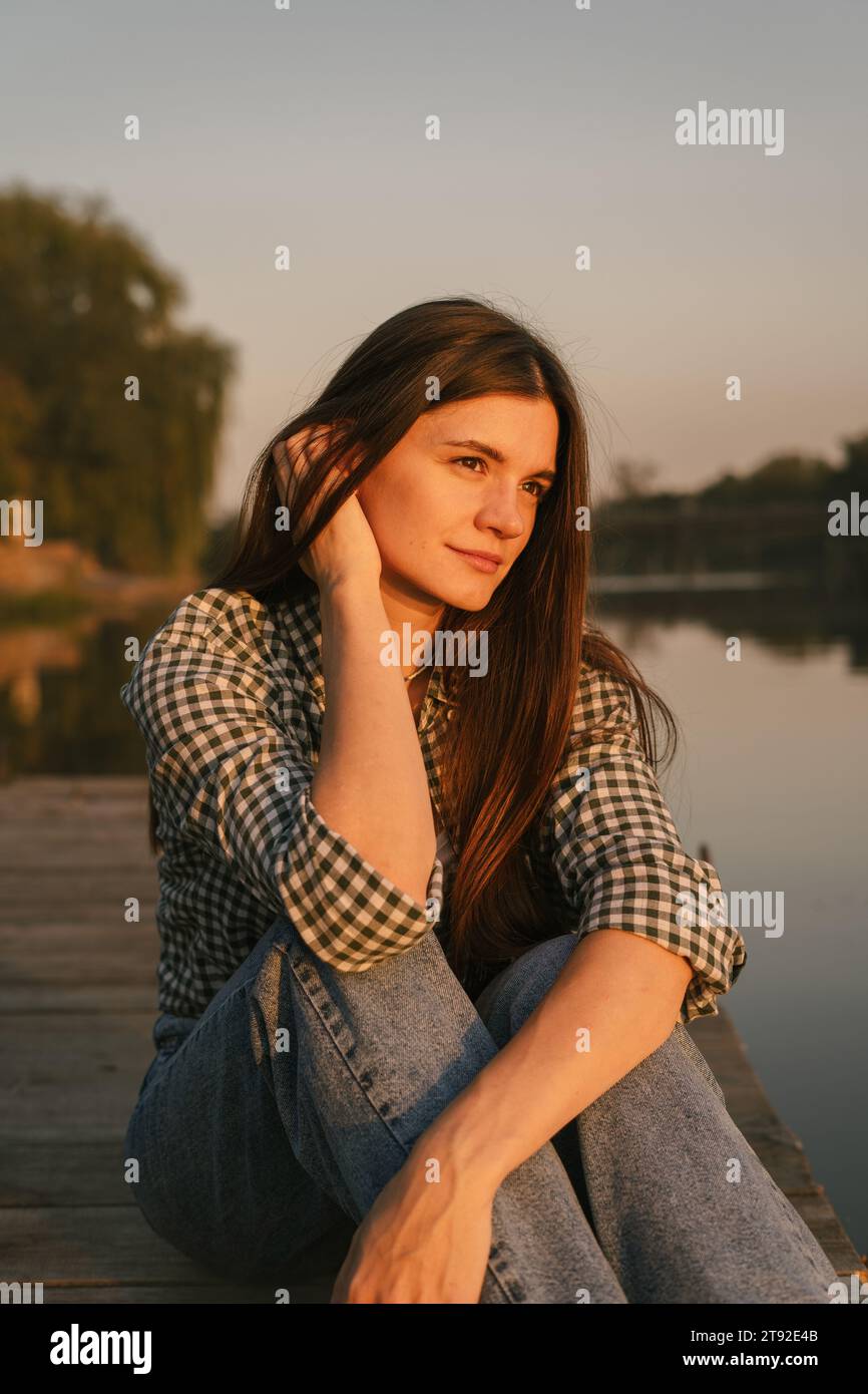 Ein Mädchen mit langen dunklen Haaren, das auf dem Flussdock sitzt. Es ist Sommer Sonnenuntergang. Das Wetter ist warm und ruhig. Stockfoto