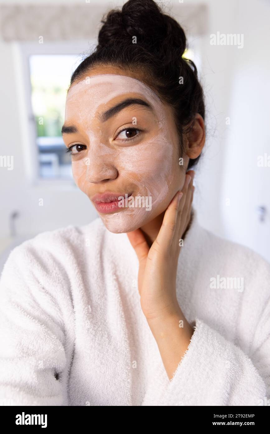Porträt einer zweirassigen Frau im Bademantel mit Gesichtsmaske im sonnigen Bad. Lebensstil, Selbstpflege, Schönheit, Haut und häusliches Leben, unverändert. Stockfoto