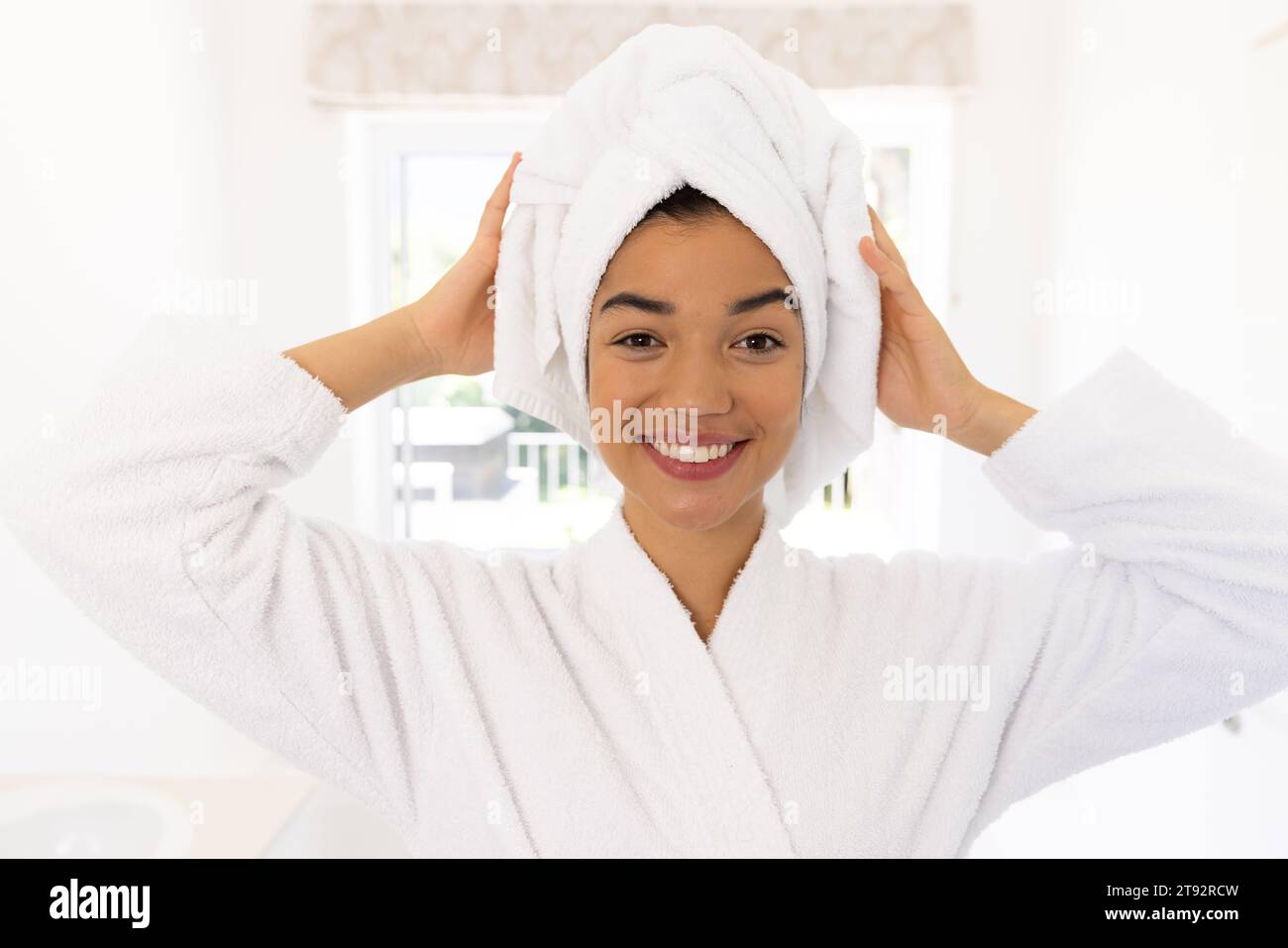 Porträt glücklicher birassischer Frau im Bademantel und mit Handtuch auf dem Kopf im sonnigen Bad. Lebensstil, Selbstversorgung und häusliches Leben unverändert. Stockfoto