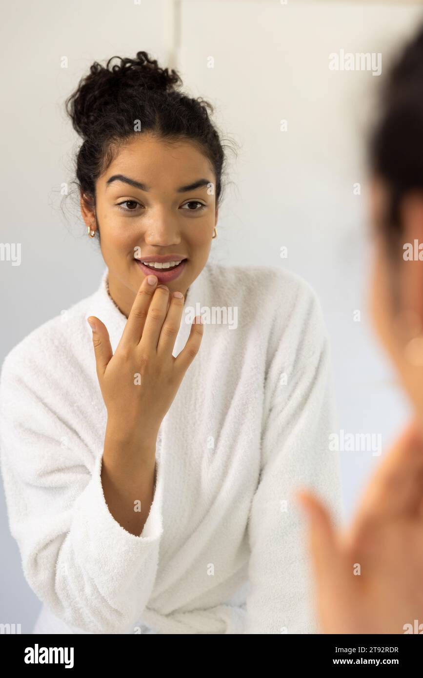 Glückliche birassische Frau im Bademantel, die im Spiegel im sonnigen Badezimmer schaut. Lebensstil, Selbstversorgung und häusliches Leben unverändert. Stockfoto
