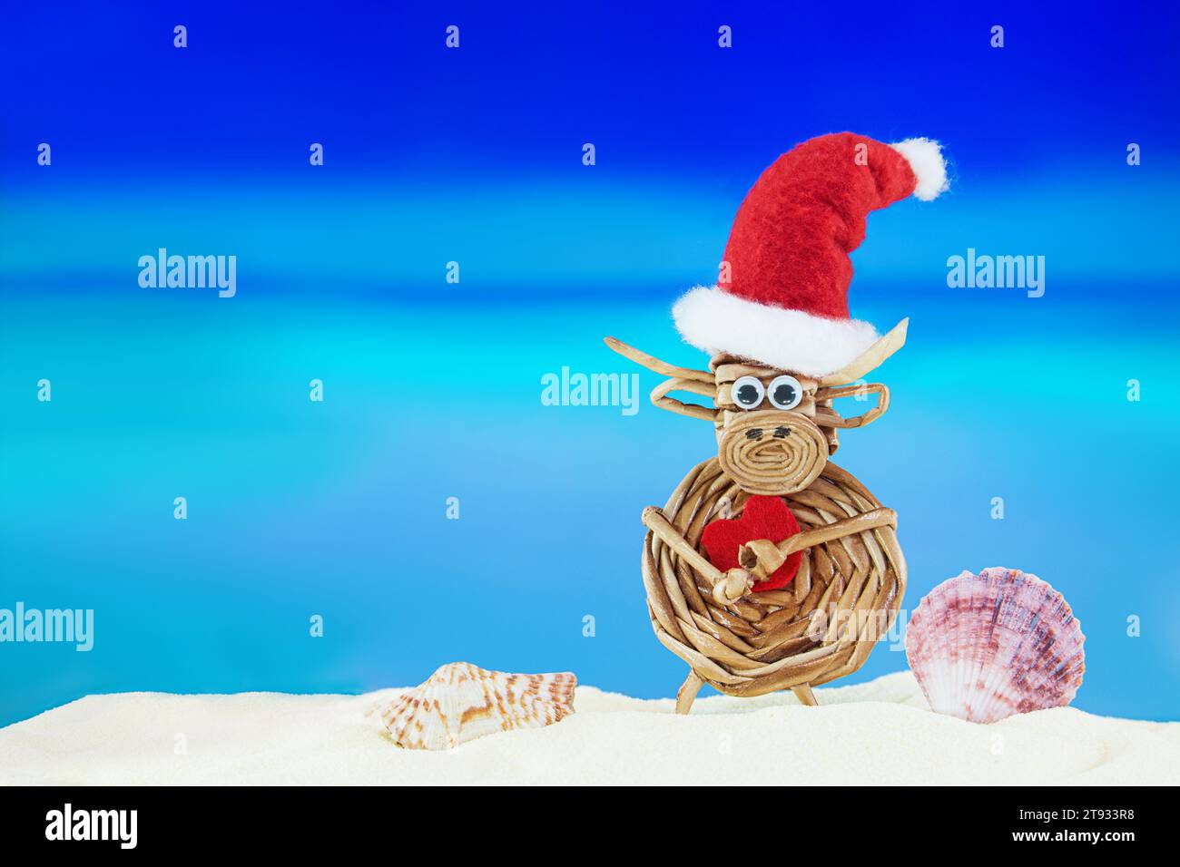 Kuh aus Spielzeuggewebe mit Weihnachtsmann-Hut mit Herz und Muscheln am Sandstrand mit Meer. Weihnachten, Neujahr, Valentinstag. Heiße Länder. Kopierbereich Stockfoto