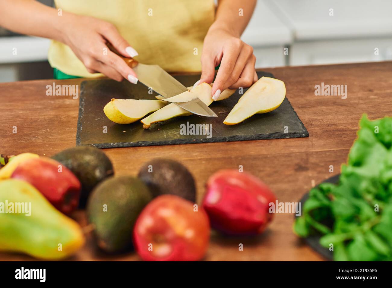 Zugeschnittene Ansicht einer vegetarischen Frau, die reife Birnen in der Nähe frischer Früchte in der Küche schneidet, pflanzliche Ernährung Stockfoto