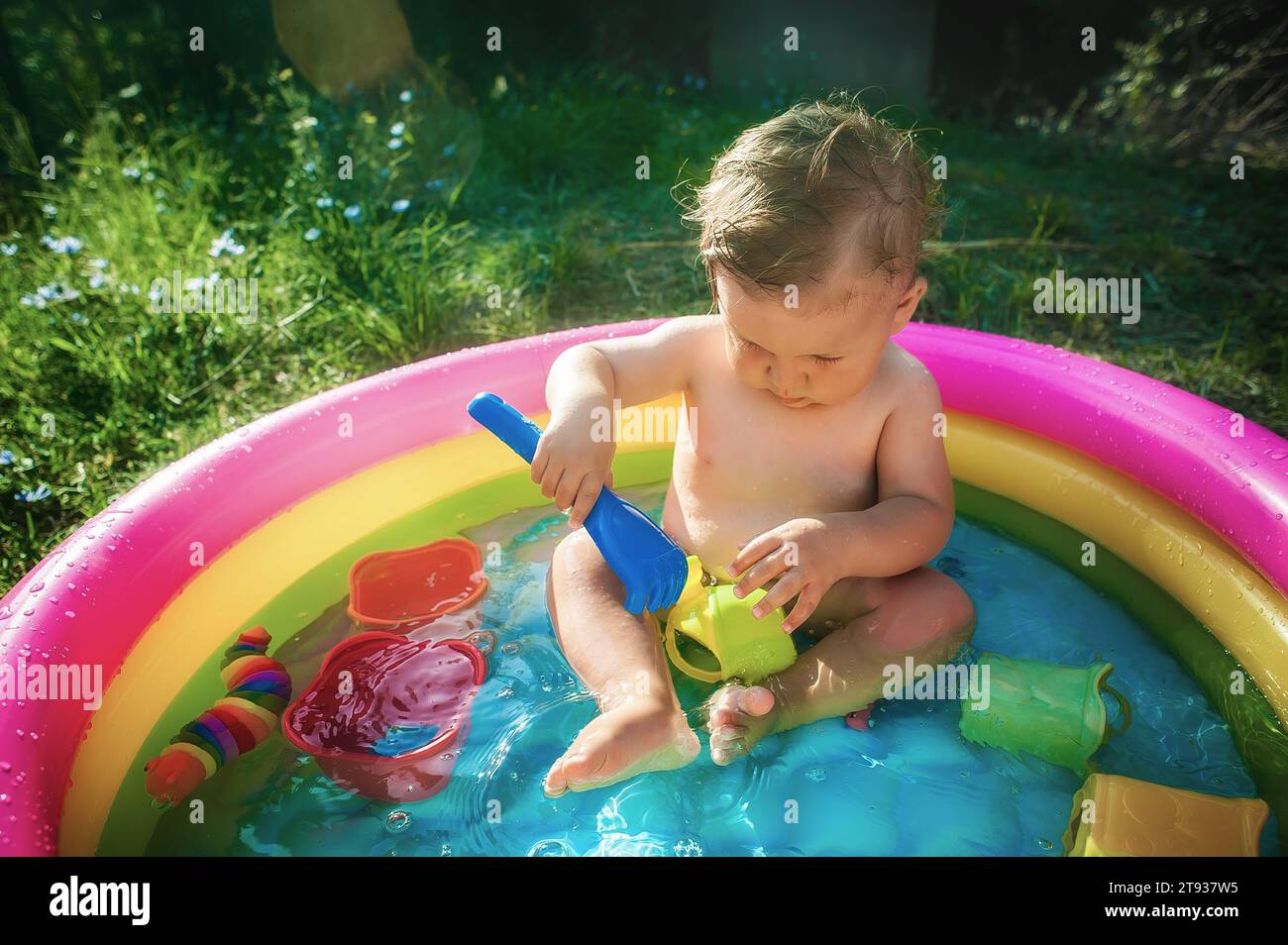 Junge, der in einem Pool im Garten sitzt und mit Wasserspielzeug spielt. Heißer Sommertag in der Natur Stockfoto