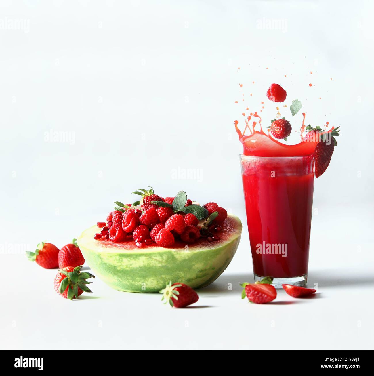 Spritzwasser mit Wassermelone, serviert mit roten Sommerfrüchten, Beeren und rotem Saft: Erdbeeren und Himbeeren fallen im Glas bei weißem Hintergrund Stockfoto