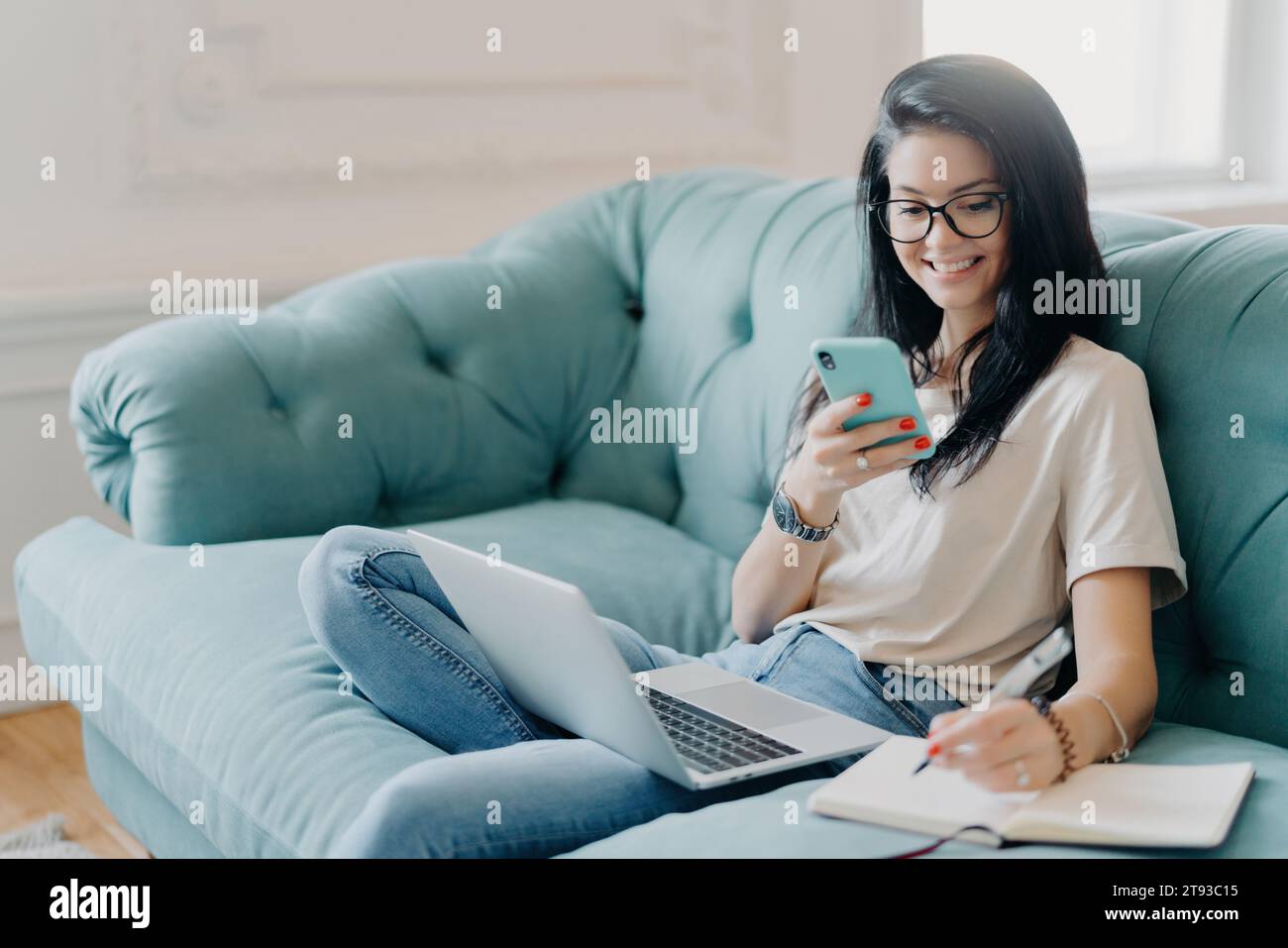 Lächelnde Schülerin, die zu Hause auf einem bequemen blauen Sofa mit Smartphone und Notebook arbeitet Stockfoto