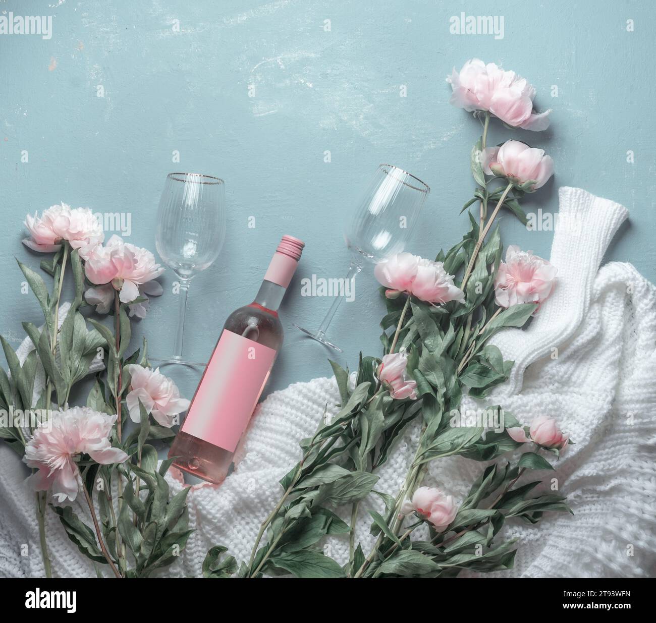 Flasche Roséwein mit Weinläsern auf blauem Hintergrund mit weißem Strickpullover und vielen rosa, blassen Pfingstrosen-Blüten. Draufsicht. Schönheit Stockfoto
