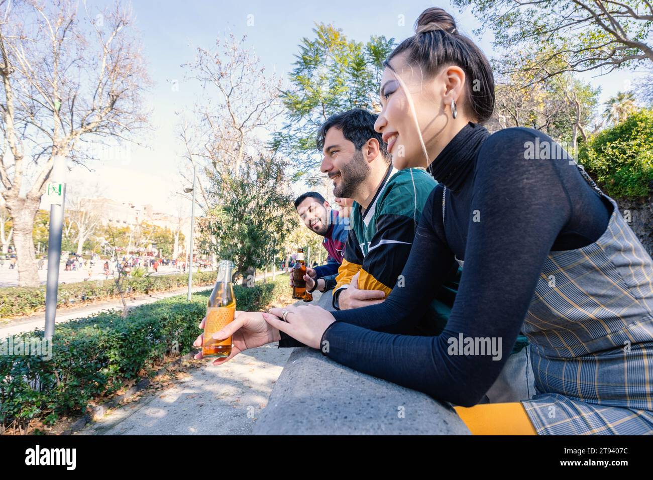 Eine vielfältige Gruppe junger Erwachsener lacht und plaudert, während sie sich an einem sonnigen Tag an einer Parkmauer ausruhen und sich Getränke teilen. Stockfoto