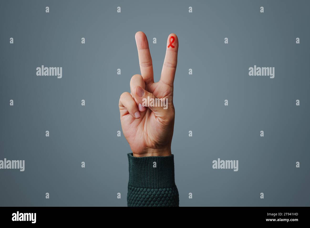 Die Hand eines Mannes, der das V-Zeichen macht, mit einem roten Wahrnehmungsband, für den Kampf gegen AIDS, in seinem Zeigefinger gemalt, vor grauem Hintergrund mit Stockfoto