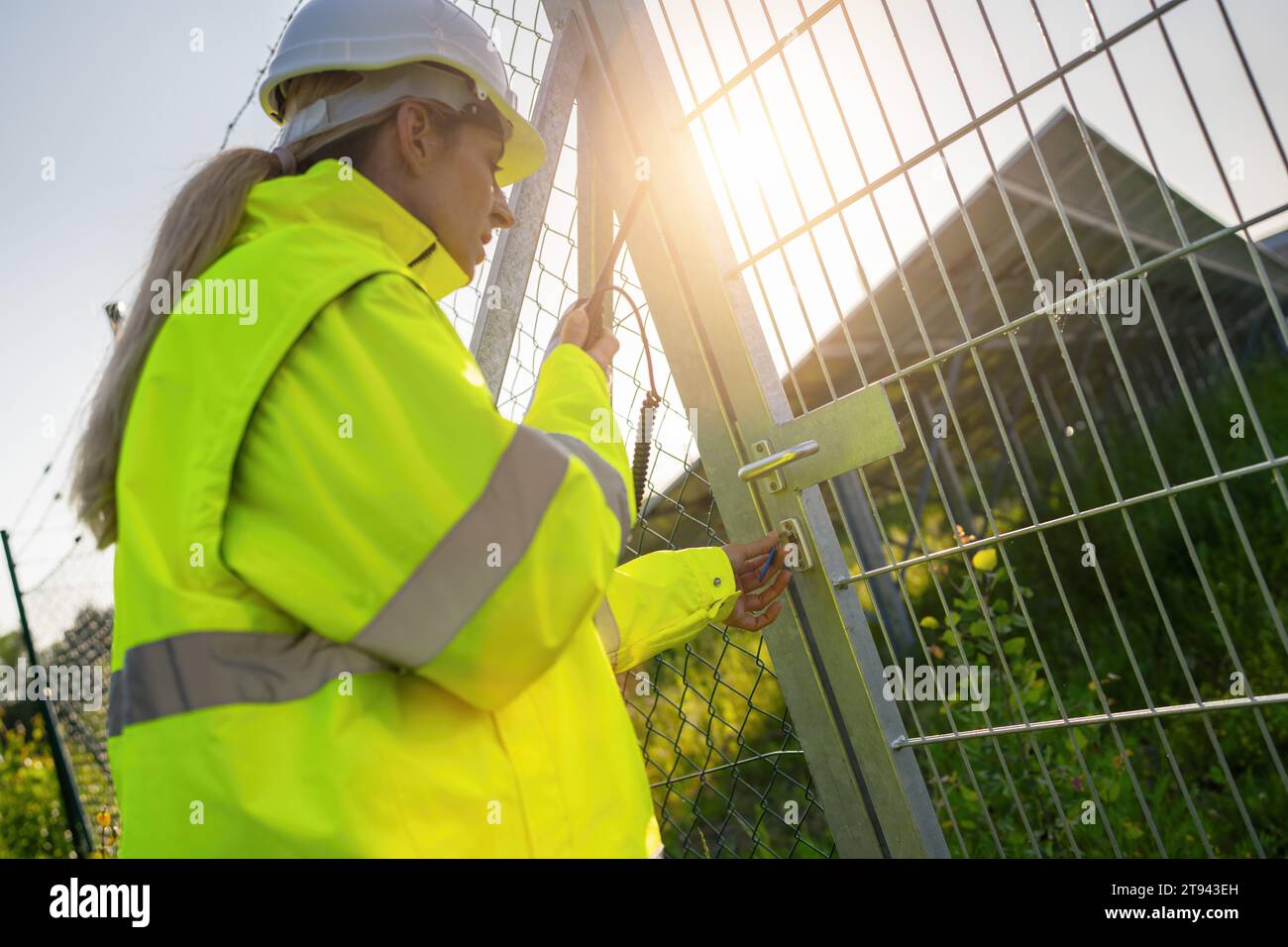 Weibliche Technikerin verriegelt das Tor in der Solaranlage während Sonnenuntergang. Alternatives energetisches ökologisches Konzeptbild. Stockfoto