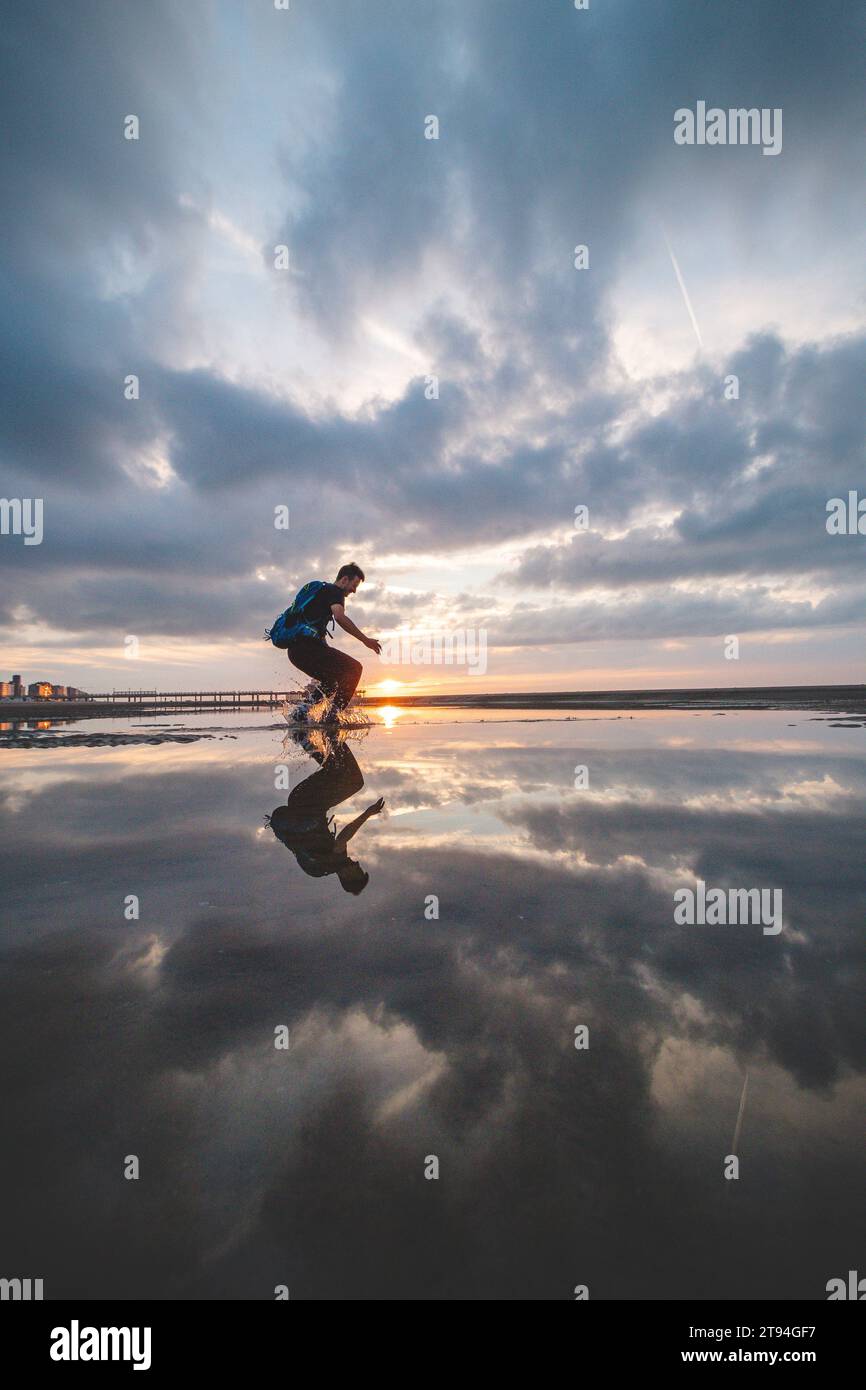 Braunhaariger Mann mit Rucksack im Alter von 25 bis 29 Jahren springt bei Sonnenuntergang in einen Wasserpool am Strand. Die Sonne zwischen dem Boden und seinen Füßen einfangen. Stockfoto