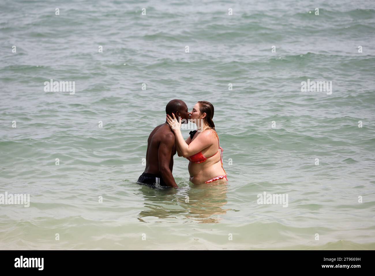 Interrassisches Paar küsst sich am Strand, weiße Frau im Bikini und Mann im Meerwasser Stockfoto