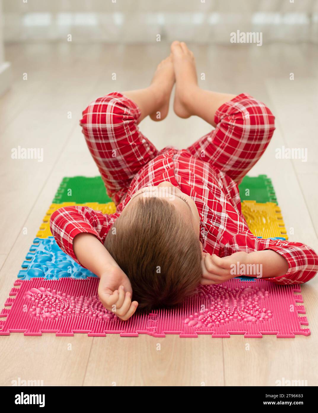 Sport- und Gesundheitskonzept. Ein kleiner Junge 4 Jahre alt im roten karierten Pyjama arbeitet auf einer mehrfarbigen orthopädischen Massage-Matte mit Stacheln in A Stockfoto