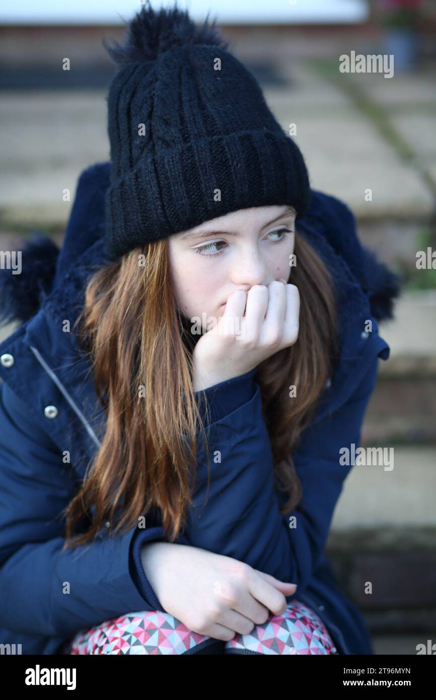 Ein Mädchen, das auf der Stufe sitzt und Wollmütze und Mantel und Schal trägt und versucht, sich warm zu halten Stockfoto