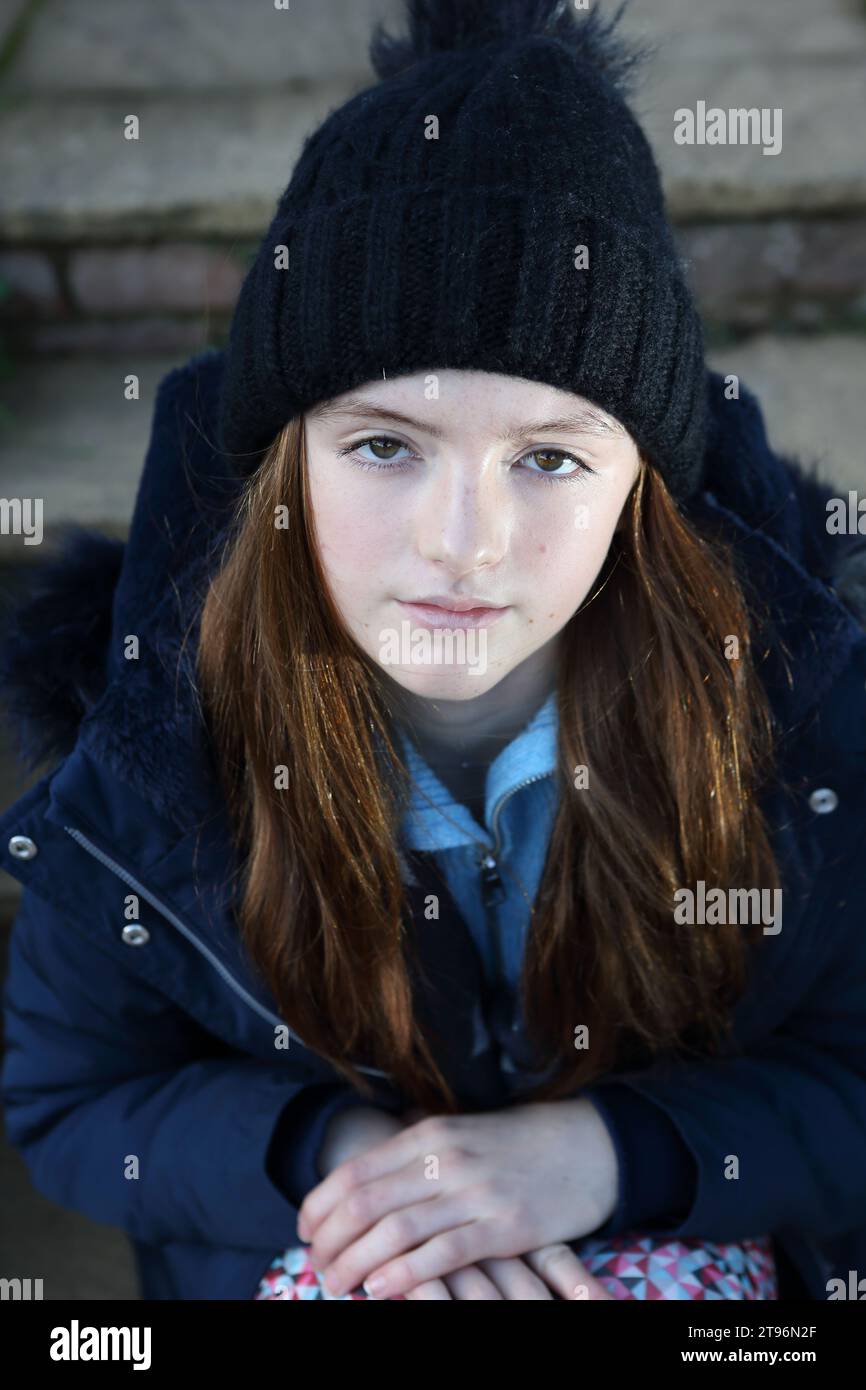 Ein Mädchen, das auf der Stufe sitzt und Wollmütze und Mantel und Schal trägt und versucht, sich warm zu halten Stockfoto