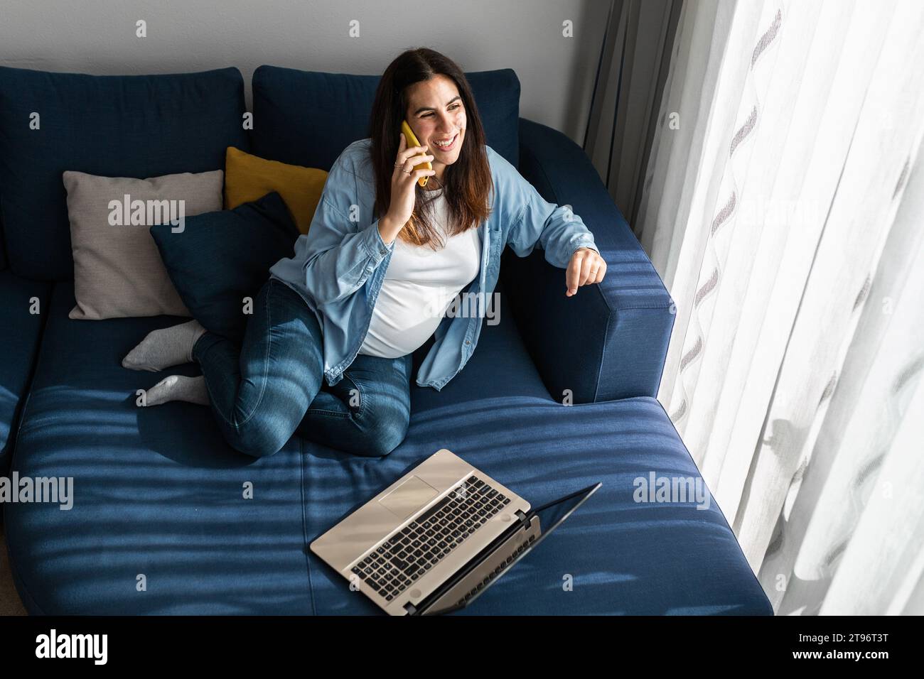 Schwangerin sitzt auf dem Sofa mit Laptop im Wohnzimmer Zimmer und sprechen auf dem Handy, während Sie von zu Hause aus arbeiten Stockfoto
