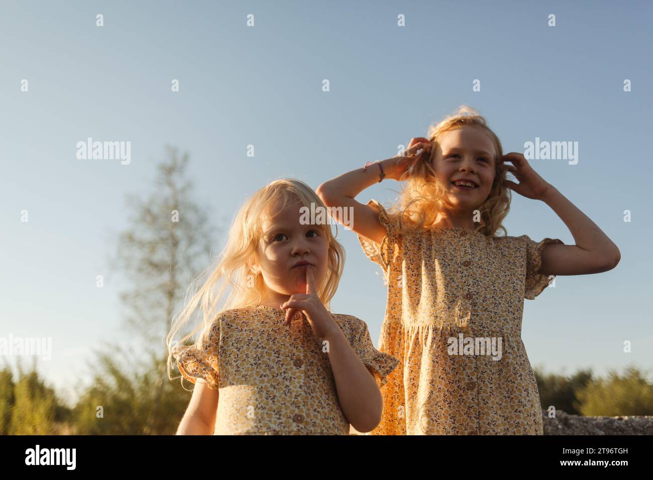 Von unten sieht ein Mädchen neugierig zu, während ihre ältere Schwester mit ihren Haaren in passenden Kleidern vor einem unscharfen Nachmittagshimmel spielt Stockfoto