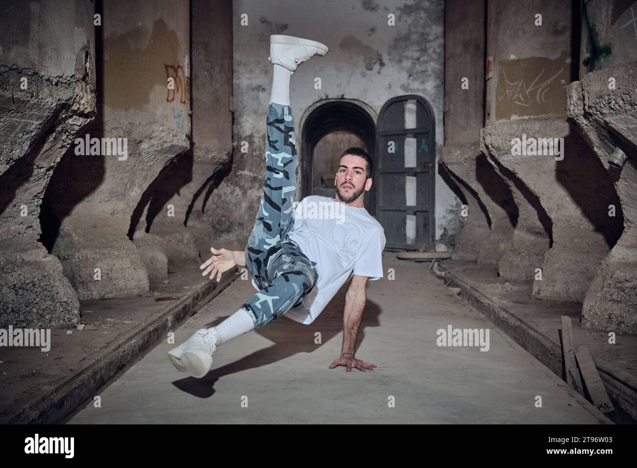 Voller Körper eines energiegeladenen bärtigen männlichen Tänzers in Freizeitkleidung, der in die Kamera schaut, während er in einem alten, verlassenen Gebäude Breakdance tanzt Stockfoto