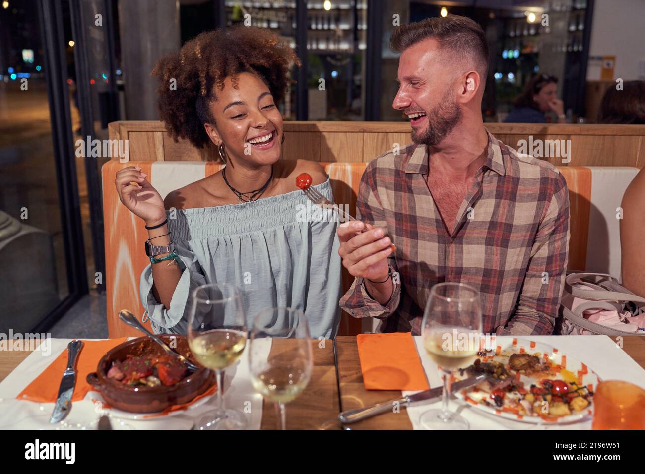 Positiver Mann, der der Afroamerikanerin Kirschtomate gibt, während er am Tisch mit Wein und leckeren Mahlzeiten im Restaurant sitzt Stockfoto
