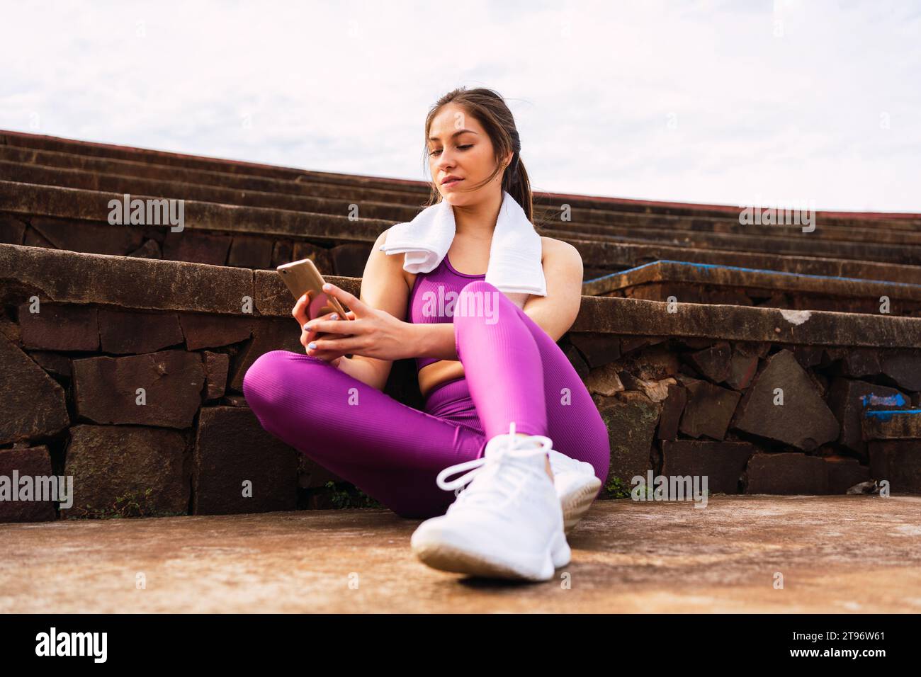 Niedriger Winkel voller Körper einer jungen weiblichen Athletin in lila Sportswear, die auf der Fußbodengrenze durch Steintreppen sitzt und an sonnigen Tagen mit Ihrem Handy herumfummelt Stockfoto