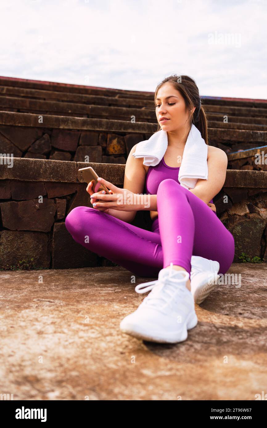Niedriger Winkel voller Körper einer jungen weiblichen Athletin in lila Sportswear, die auf der Fußbodengrenze durch Steintreppen sitzt und an sonnigen Tagen mit Ihrem Handy herumfummelt Stockfoto