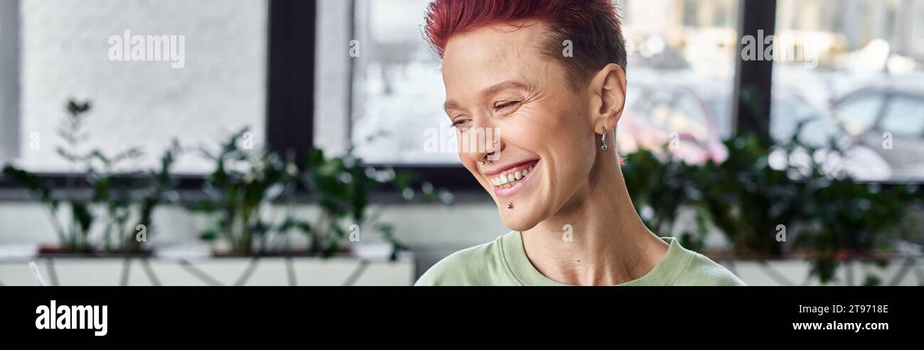 Stilvolle und fröhliche nicht-binäre Person mit kurzen Haaren, die im Büro lachen, horizontales Banner Stockfoto