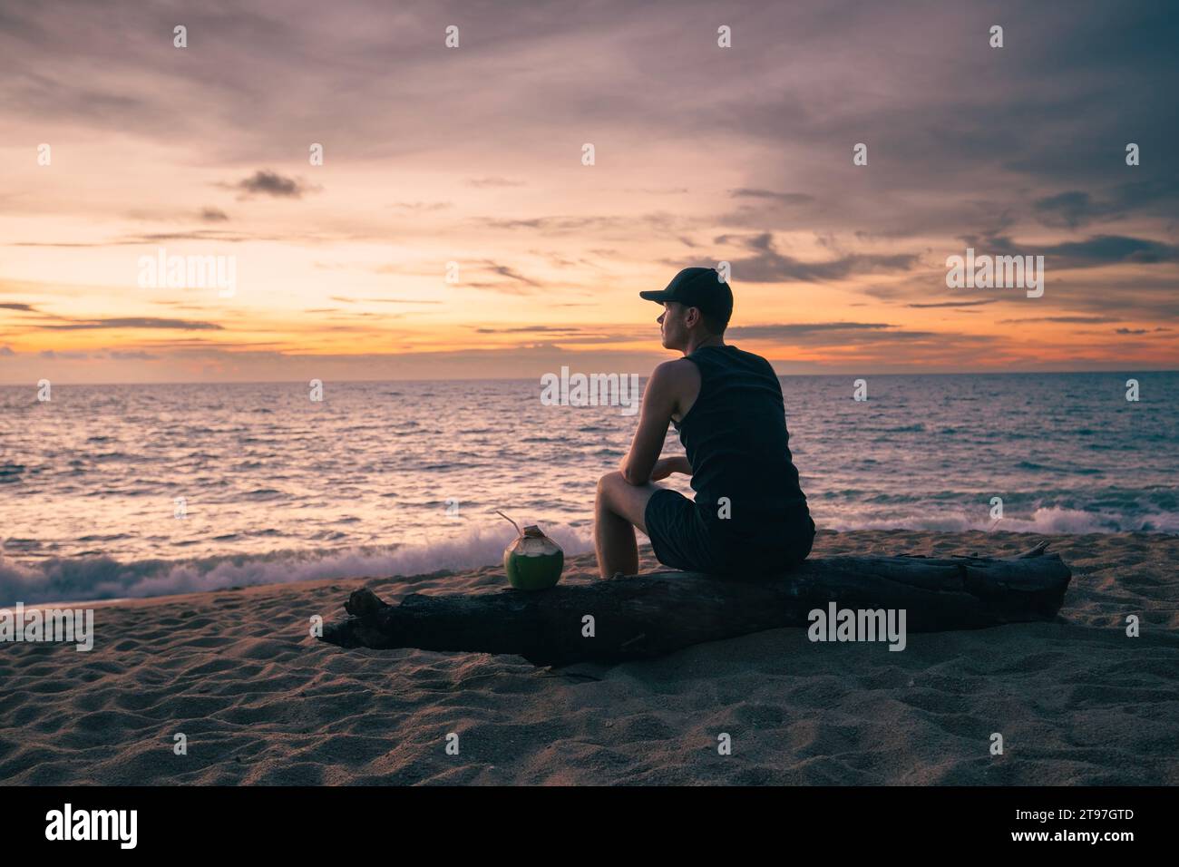 Mann sitzt am tropischen Sandstrand mit Kokosnussgetränk und genießt den farbenfrohen Sonnenuntergang über dem Meer. Themen für Alleinreisende und Buget-Reisen. Stockfoto