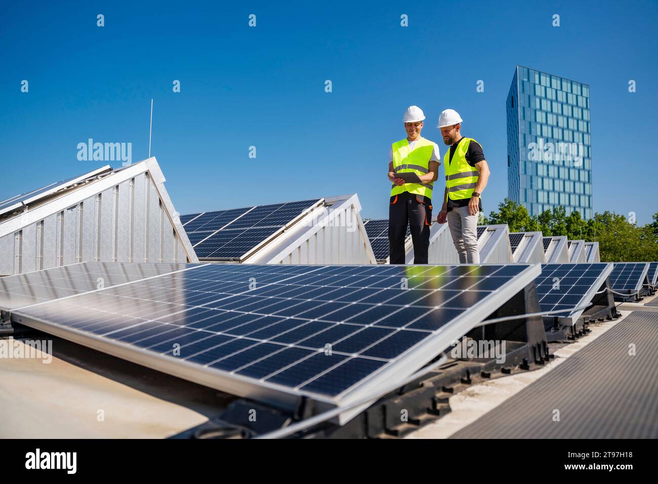 Zwei Techniker, die einen Tablet-PC verwenden, während sie auf dem Dach eines Firmengebäudes arbeiten, das mit Solarpaneelen ausgestattet ist Stockfoto