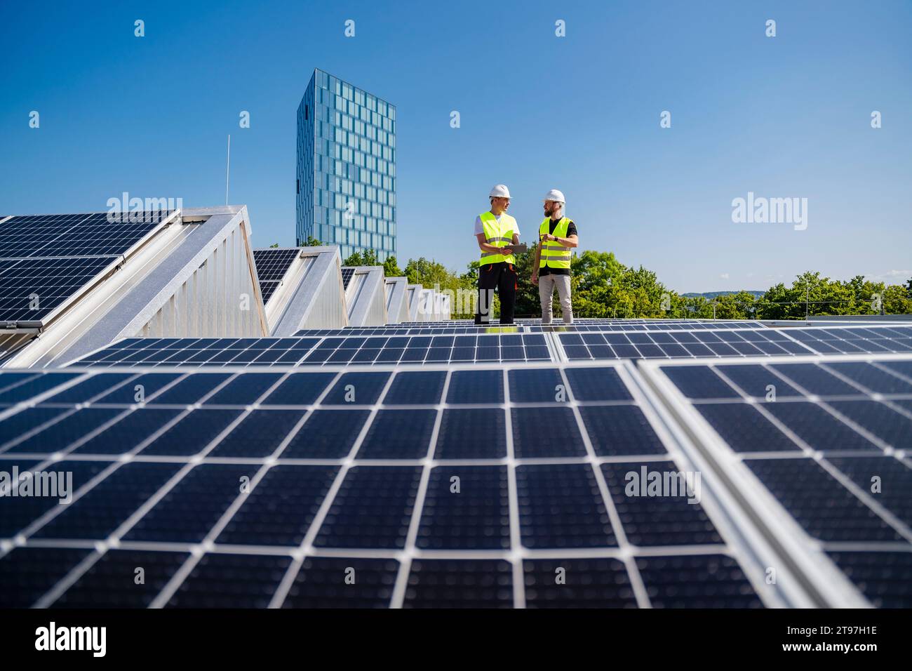 Zwei Techniker, die einen Tablet-PC bei der Inspektion von Solarpaneelen auf dem Dach eines Firmengebäudes verwenden Stockfoto