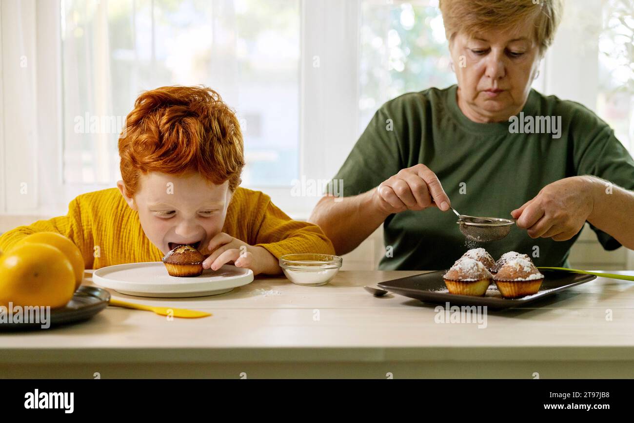 Großmutter staubt Zucker auf Cupcakes mit Enkel, der zu Hause in der Küche isst Stockfoto
