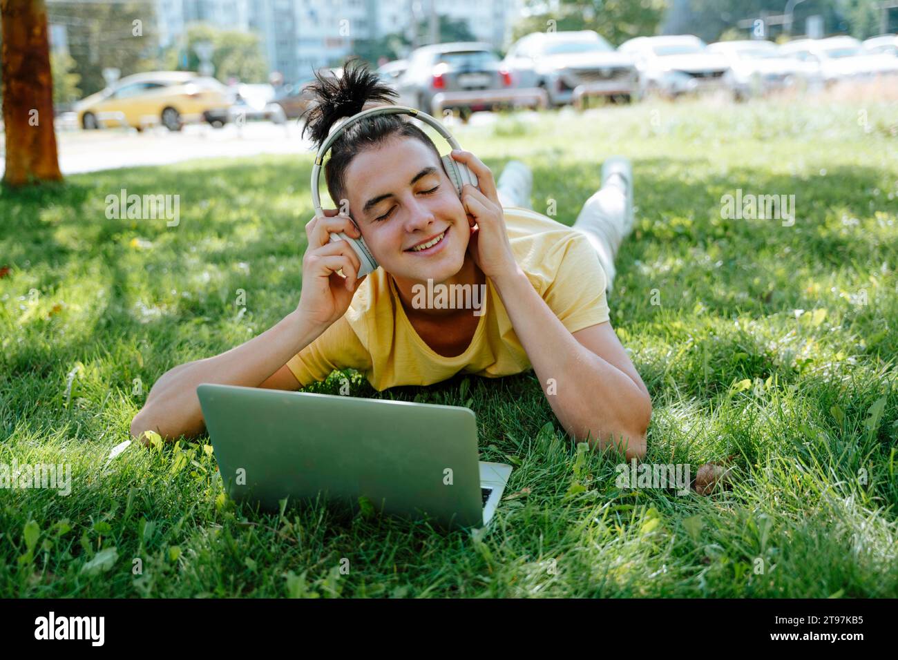 Lächelnder Mann mit kabellosen Kopfhörern, der im Gras liegt und mit dem Laptop im Park liegt Stockfoto