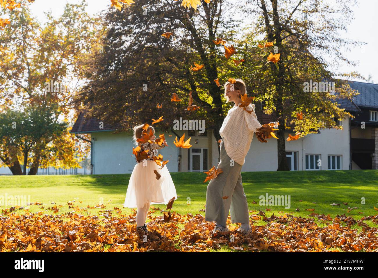 Glückliche Mutter und Tochter spielen mit Herbstlaub vor dem Haus Stockfoto
